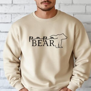 May include: A beige crewneck sweatshirt with the words "papa BEAR" in black script and a bear silhouette. The sweatshirt is made of a soft material and is suitable for casual wear.