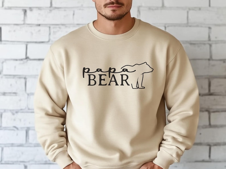 May include: A beige crewneck sweatshirt with the words "papa BEAR" in black script and a bear silhouette. The sweatshirt is made of a soft material and is suitable for casual wear.