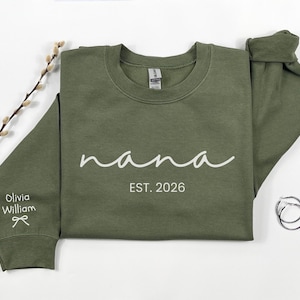 May include: An olive green sweatshirt with the word "nana" in white script, and "EST. 2026" below. The sleeve has the name "Olivia William" and a bow. Silver hoop earrings are on the right.