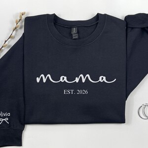 以下が含まれることがあります： 白の筆記体で「mama」の文字と「EST. 2026」の年号がプリントされたネイビーブルーのスウェットシャツ。袖口には「Olivia」の名前とリボンが描かれています。シルバーのフープピアスが見えます。
