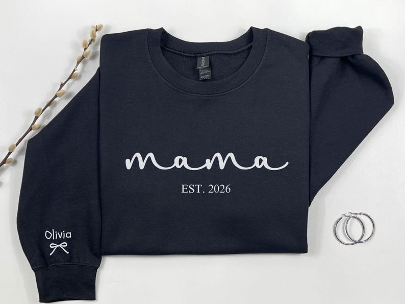 以下が含まれることがあります： 白の筆記体で「mama」の文字と「EST. 2026」の年号がプリントされたネイビーブルーのスウェットシャツ。袖口には「Olivia」の名前とリボンが描かれています。シルバーのフープピアスが見えます。
