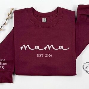 以下が含まれることがあります： 白い筆記体で「mama」という文字と「EST. 2026」の年号が入ったマルーン色のスウェットシャツ。左袖には「Olivia William」の名前とリボンのグラフィック。右側にはシルバーのフープピアスがあります。