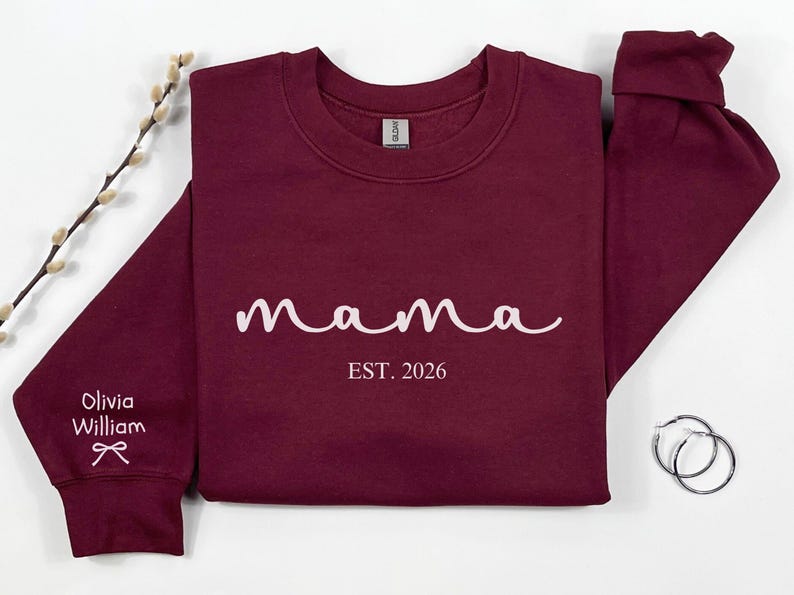 以下が含まれることがあります： 白い筆記体で「mama」という文字と「EST. 2026」の年号が入ったマルーン色のスウェットシャツ。左袖には「Olivia William」の名前とリボンのグラフィック。右側にはシルバーのフープピアスがあります。