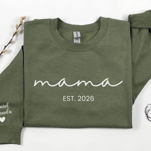 May include: An olive green sweatshirt with the word "mama" in white script, and the text "EST. 2026". The left sleeve has the names "Daniel Amanda" and a heart. A sprig of pussy willow and silver hoop earrings are also in the image.