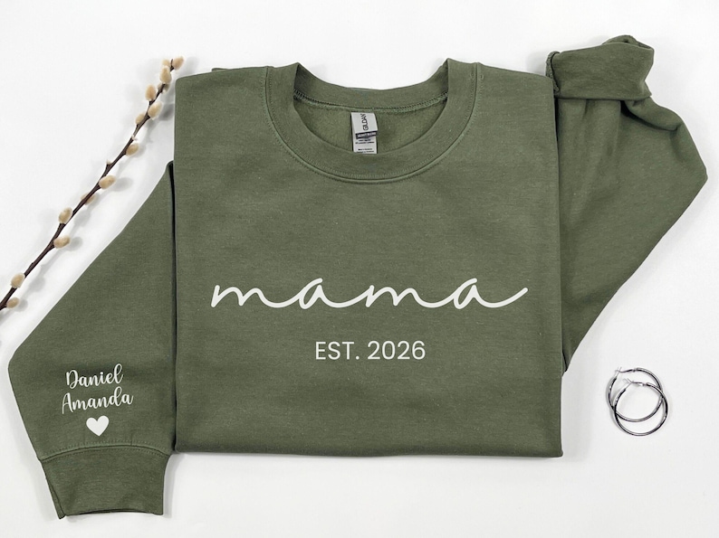 May include: An olive green sweatshirt with the word "mama" in white script, and the text "EST. 2026". The left sleeve has the names "Daniel Amanda" and a heart. A sprig of pussy willow and silver hoop earrings are also in the image.
