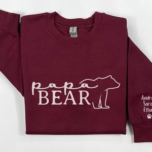 May include: A maroon sweatshirt with the words "papa BEAR" in white script and a bear outline. The right sleeve has the names "Andrew Sarah Ethan" and a paw print in white.
