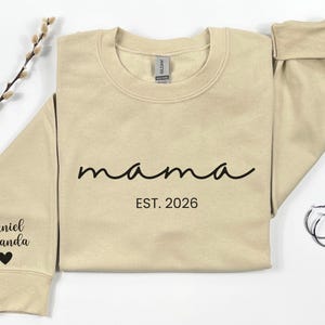 May include: A beige sweatshirt with the word "mama" in black script, and "EST. 2026" printed below. The left sleeve has the names "Daniel Amanda" and a heart. A pair of silver hoop earrings are on the right.