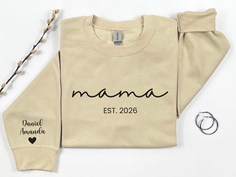 May include: A beige sweatshirt with the word "mama" in black script, and "EST. 2026" printed below. The left sleeve has the names "Daniel Amanda" and a heart. A pair of silver hoop earrings are on the right.