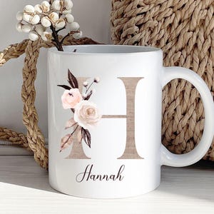 May include: White ceramic mug featuring a brown letter H and a floral design. The mug is personalised with the name "Hannah" beneath the letter.