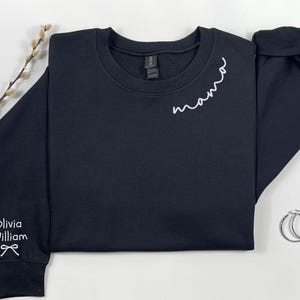 May include: A navy blue sweatshirt with the word "mama" embroidered in white script on the chest. The left sleeve cuff has the name "Olivia William" embroidered in white. Silver hoop earrings are next to the sweatshirt.