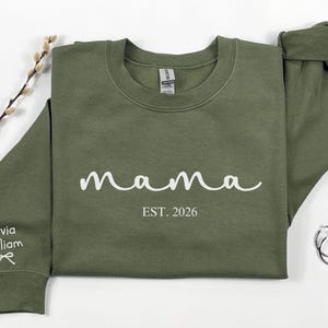 以下が含まれることがあります： 白い筆記体で「mama」の文字と「EST. 2026」の文字が入ったオリーブグリーンのスウェットシャツ。左袖には「Olivia William」という名前とリボンのグラフィックが入っています。シルバーのフープイヤリングとヤナギの枝も画像にあります。