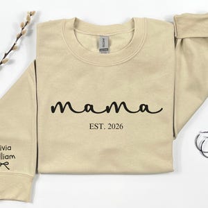 以下が含まれることがあります： 黒い筆記体で「mama」と書かれたベージュのスウェットシャツ、その下に「EST. 2026」の文字。左袖には「Olivia William」という名前とリボンのグラフィック。右側にはシルバーのフープピアスがあります。