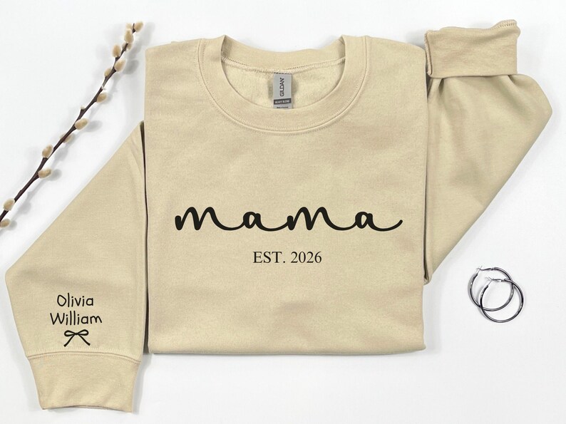 以下が含まれることがあります： 黒い筆記体で「mama」と書かれたベージュのスウェットシャツ、その下に「EST. 2026」の文字。左袖には「Olivia William」という名前とリボンのグラフィック。右側にはシルバーのフープピアスがあります。