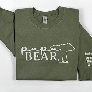 May include: Olive green sweatshirt with the words "Papa Bear" and a bear graphic in white. The sleeve has the names "Andrew, Sarah, Ethan" and a paw print.