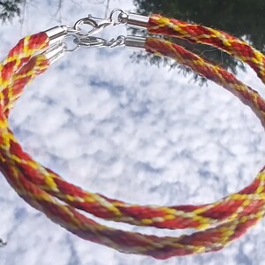 May include: Two red, yellow, and white braided cords with silver clasps. The cords are twisted together to form a decorative bracelet.