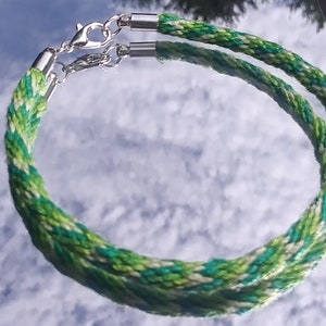 Puede incluir: Un brazalete de cordón trenzado verde, blanco y turquesa con cierres plateados.