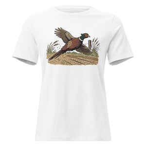 May include: White t-shirt featuring a detailed illustration of a pheasant in flight. The bird is depicted with brown, black, and red feathers, set against a backdrop of a fence, wheat, and a dirt path. The design is printed on a classic crew neck tee.