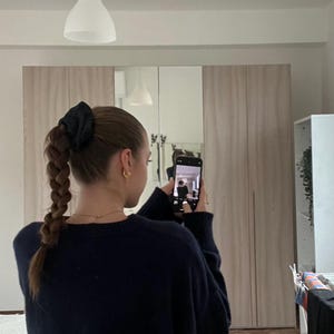 May include: A person with long brown hair pulled back in a braid with a black scrunchie takes a selfie in a mirror. They are wearing a dark blue sweater.
