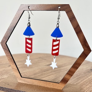 May include: A pair of patriotic earrings featuring a blue triangle top, a red rectangular body with white stripes, and a white star at the bottom. The earrings are suspended from silver-colored fish hook ear wires.