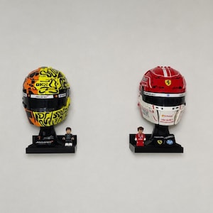 May include: Two buildable racing helmet models on black display stands. The left helmet is yellow and orange with black patterns and McLaren branding. The right helmet is red and white with Ferrari logos. Each includes a small matching minifigure.