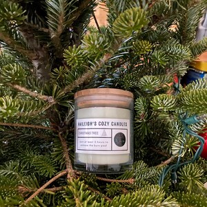 Op de afbeelding: Een glazen kaarsenpot met een houten deksel, genesteld tussen groenblijvende takken. Het etiket luidt "Haileigh's Cozy Candles" en "Christmas Tree". De kaars is lichtgroen van kleur. Het etiket bevat ook de tekst "Minimaal 2 uur branden om de brandpoel te bereiken."