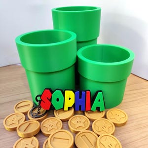 May include: A collection of green pipe-shaped containers, a keychain with the name "SOPHIA" in colorful letters, and several gold-colored coin-shaped objects. The items are arranged on a light brown surface.
