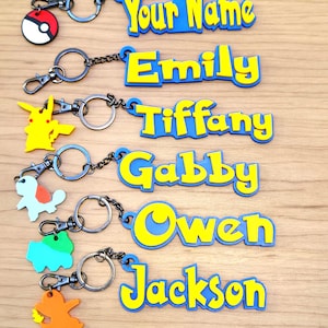May include: Keychains with personalized names and cartoon character charms. The names are in yellow, outlined in blue. Charms include a Pokéball, Pikachu, Squirtle, Bulbasaur, and Charmander. Each keychain has a silver chain and clasp.