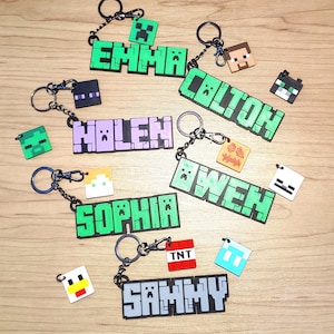May include: A collection of personalized keychains with names in pixelated lettering, reminiscent of a popular video game. Each keychain features a name in a different color, with matching character charms. The keychains are attached to metal rings.