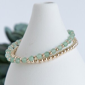 May include: Two delicate bracelets. One features small, round, light green beads, and the other has small, gold-coloured beads. The bracelets are stacked on a white, cone-shaped display, with a blurred green plant in the background.