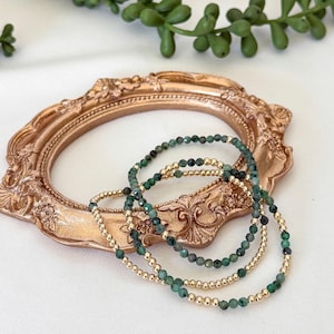 Op de afbeelding: Drie armbanden met smaragdgroene en gouden kralen worden getoond op een goudkleurige, sierlijke fotolijst. De armbanden zijn gelaagd en tonen het contrast tussen de groene en gouden kleuren. Een takje groen loof staat op de achtergrond.