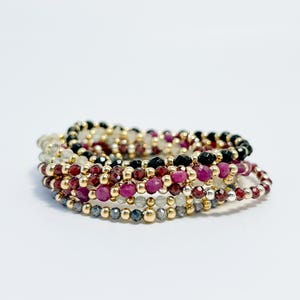 Puede incluir: Un conjunto de pulseras de cuentas en varios colores, incluyendo rojo, negro y transparente, con cuentas doradas. Las pulseras están dispuestas en forma circular, mostrando los diferentes colores y texturas de las piedras preciosas.