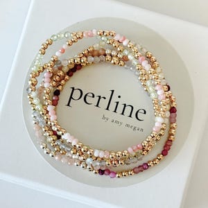 Puede incluir: Un conjunto de delicados brazaletes de cuentas doradas con cuentas de piedras preciosas rosas, verdes y marrones. Los brazaletes están apilados uno encima del otro y se encuentran en una caja blanca con el texto "perline by amy megan".