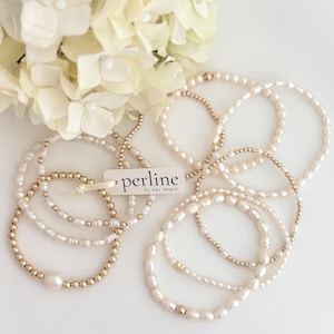 Puede incluir: Varios brazaletes de perlas y cuentas doradas están dispuestos sobre una superficie blanca, con un ramo de flores blancas en el fondo. Una etiqueta con la palabra "perline" está adherida a uno de los brazaletes.