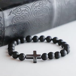 Puede incluir: Pulsera de cuentas negras con un colgante de cruz. La pulsera está hecha de cuentas redondas negras mate y una cruz gris oscuro. La pulsera se muestra sobre una superficie blanca con un fondo oscuro texturizado.