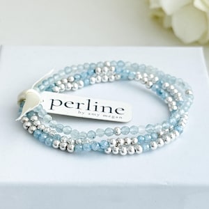May include: Three delicate bracelets with light blue and silver beads. The bracelets are stacked together and are on a white surface. A tag with the text "perline by amy megan" is attached to the bracelets.