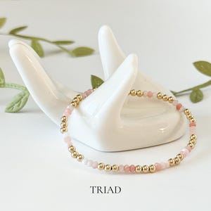 Puede incluir: Un delicado brazalete hecho con peque&ntilde;as cuentas doradas y cuentas de piedras preciosas rosas y blancas. El brazalete se muestra en una mano de cer&aacute;mica blanca.
