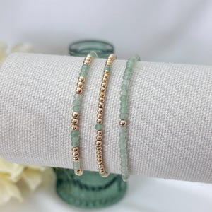 May include: A set of three beaded bracelets featuring alternating small round gold-toned metallic beads and faceted light green gemstone beads. The bracelets are arranged side-by-side, showcasing a delicate design with consistent bead sizing.