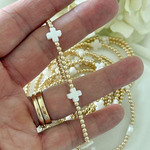 May include: Gold beaded necklace with white cross charms. The necklace features small gold beads and white cross-shaped pendants. The design is elegant and suitable for religious or fashion purposes.