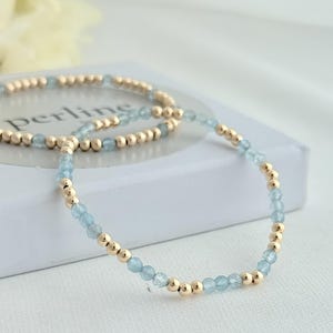 Puede incluir: Dos pulseras delicadas. Cada pulsera presenta cuentas azul claro y doradas alternadas. Las pulseras se exhiben sobre una superficie blanca. La palabra "Perline" es parcialmente visible en la superficie.