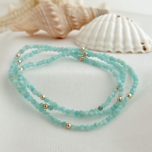 May include: A delicate, layered bracelet featuring small, light blue beads and gold-colored accents. The bracelet is displayed on a white surface, with seashells in the background. The jewelry has a simple, elegant design.