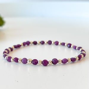 May include: A purple gemstone bracelet with gold beads. The bracelet is made with small, oval-shaped gemstones and gold beads.