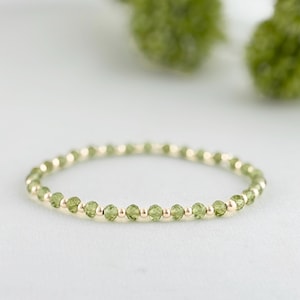 May include: A delicate gold ring featuring small, round, light green peridot gemstones interspersed with tiny gold beads. The ring is set against a soft white background with blurred green accents.