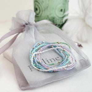 May include: Several delicate beaded bracelets in pastel shades of blue, green, and lilac, with gold-coloured beads. The bracelets are presented on a grey organza bag with the word "line" printed on it.