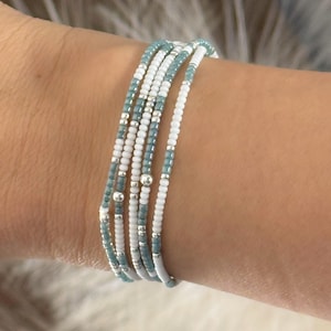 Puede incluir: Un conjunto de pulseras de cuentas blancas, azul claro y plateadas. Las pulseras están hechas de pequeñas cuentas ensartadas, creando un aspecto delicado y en capas. Las pulseras se llevan en la muñeca.