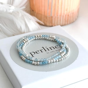 May include: Three delicate bracelets with light blue and silver beads are displayed on a white box. The bracelets are stacked together, creating a layered effect. The box has the text "perline by amy mega" printed on it.