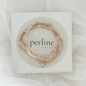 May include: Two beaded bracelets with rose quartz and gold-colored beads, presented in a white box. The bracelets are layered, with a heart-shaped charm on the lower bracelet. The box has the text "perline by amy megan" in a circular design.