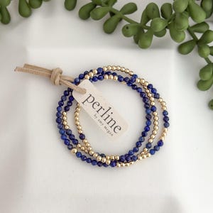 Op de afbeelding: Drie armbanden met kralen. Twee armbanden hebben kleine, donkerblauwe kralen en de derde armband heeft kleine, goudkleurige kralen. Een label met het woord "perline" is met een leren koord aan de armbanden bevestigd. De armbanden liggen op een witte ondergrond.