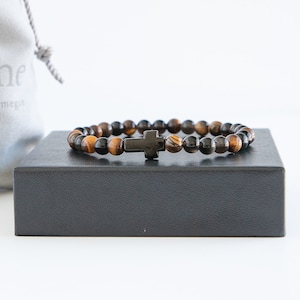 May include: A brown and black beaded bracelet with a black cross charm. The bracelet is on a black box.