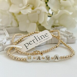 Könnte beinhalten: Zwei goldfarbene Perlenarmbänder, eines mit weißen Buchstabenperlen, die "MAMA" buchstabieren. Das andere ist ein schlichtes goldfarbenes Perlenarmband. Ein Etikett trägt die Aufschrift "perline by amy megan". Der Hintergrund zeigt weiße Blumen.