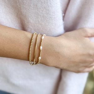 May include: Three gold bracelets are stacked on a wrist. One bracelet is made of small gold beads, another has a gold bar, and the third features small gold beads interspersed with white pearls. The bracelets are worn on a person's wrist.